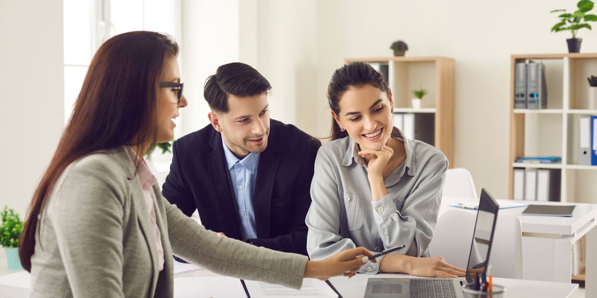 Three people having a business meeting, looking at a laptop screen, with one person pointing at the screen."